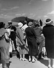 Crowd stands behind a fence, looking onto the airfield at an airpl- Old Photo